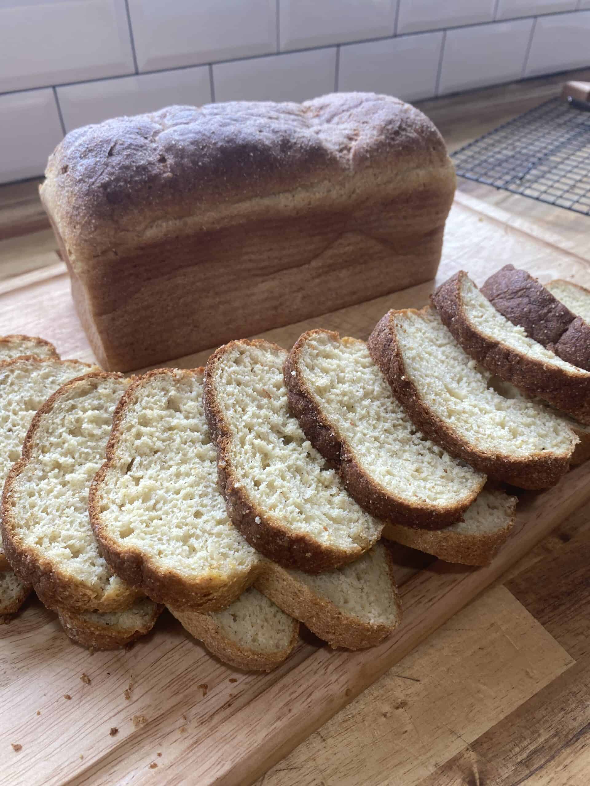 Gluten free fresh milled sandwich bread loaves positioned on a wooden cutting board for a photo with subway tile in the background and a black cooling rack in the background. One loaf is fully in tact standing on the board and the other loaf has been sliced with 9 slices visible. The gluten free bread has a great crumb and browned edges that have been sifted by buttering after removing from the oven. Miscellaneous crumbs scatter around the bread on the wooden board.