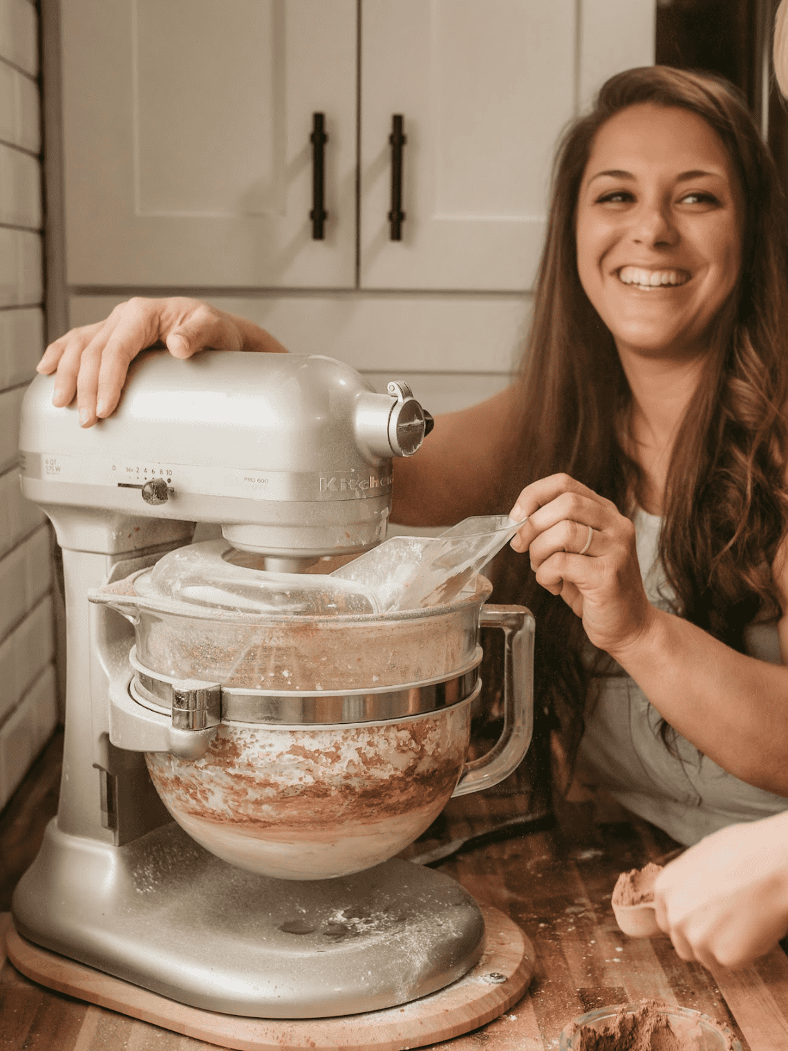 Cookbook author, Jennifer Roberts, is mixing chocolate icing. Her son is holding a scoop of cocoa powder over a messy counter. The mixer is a silver kitchen aid professional with a glass bowl. Jennifer is smiling showing her teeth at her son as she holds onto the machine while it mixes. She is wearing light blue overalls with a white t-shirt underneath. There is a knife on the countertop. The countertop is wood grains (acacia). The tile on the wall is subway tile and Jennifer has a white cabinet with black handles behind her.