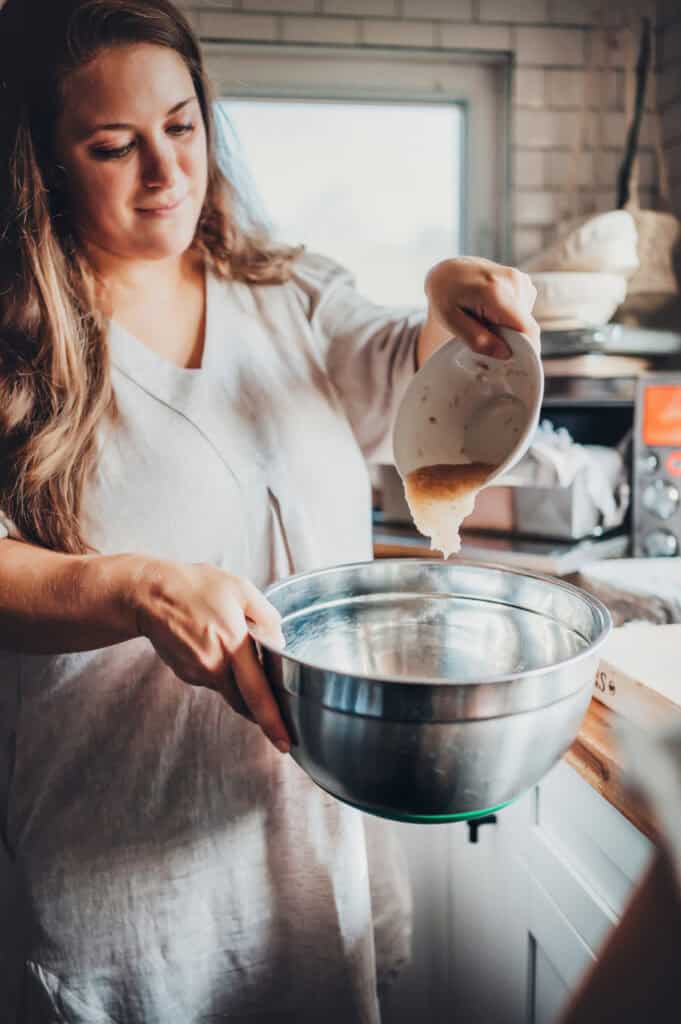 Jennifer Roberts is pouring psyllium husk gel into a bowl wearing a tan dress. In the background there is a countertop oven with sourdough containers on top, a window, and white subway backsplash.