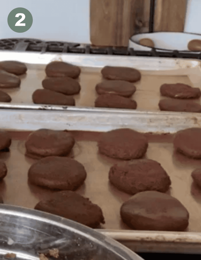 Two rectangular metal baking sheets with 4 rows of circle dark brown cookies spaced evenly and a small green circle with a white number two in the upper left hand corner
