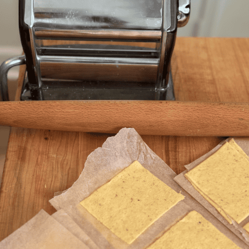 Square wonton wrappers on parchment paper with a rolling pin and pasta roller.