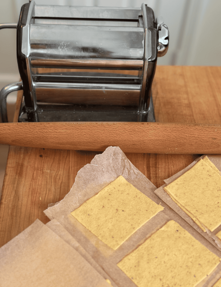 Square wonton wrappers on parchment paper with a rolling pin and pasta roller.