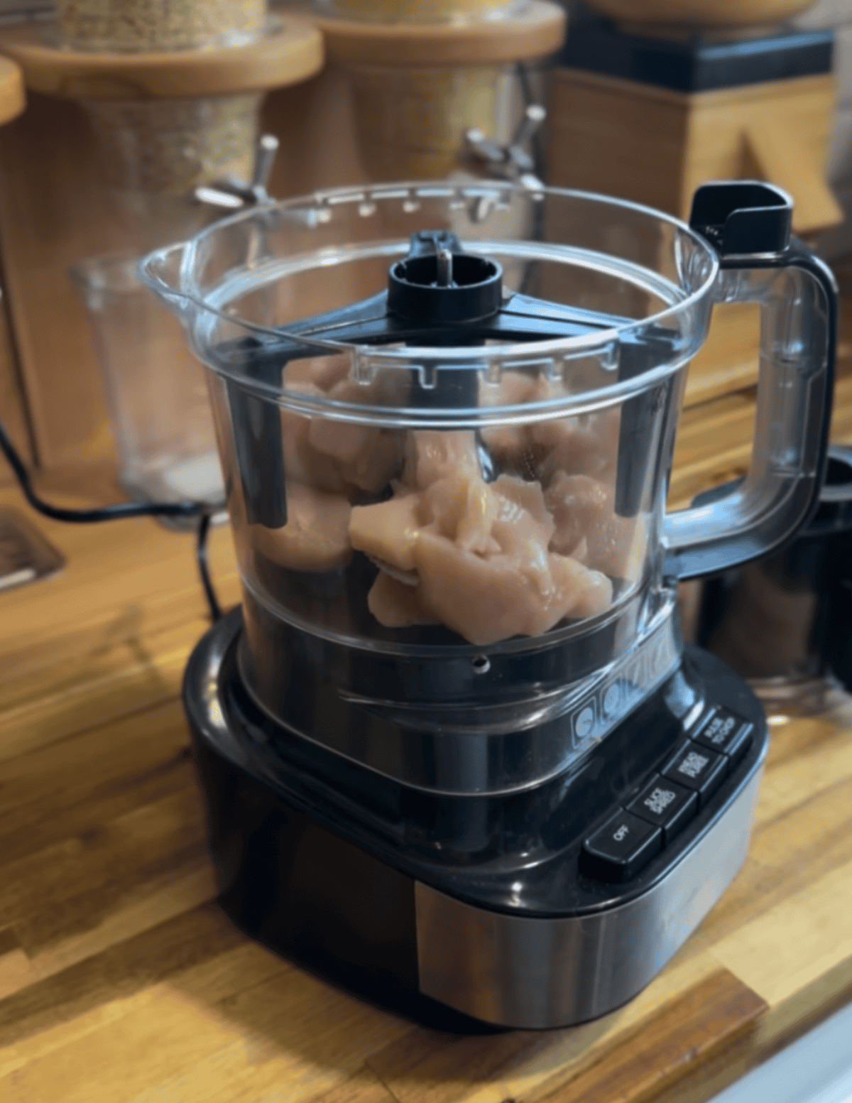Food processor on the counter with chunks of chicken and seasoning inside.