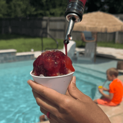 Pouring a mixed berry dye-free and refined sugar free snow cone syrup over shaved ice in a white snow cone cup. There is a little boy in an orange bathing suit and a pool in the background.