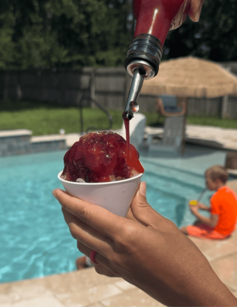 Pouring a mixed berry dye-free and refined sugar free snow cone syrup over shaved ice in a white snow cone cup. There is a little boy in an orange bathing suit and a pool in the background.