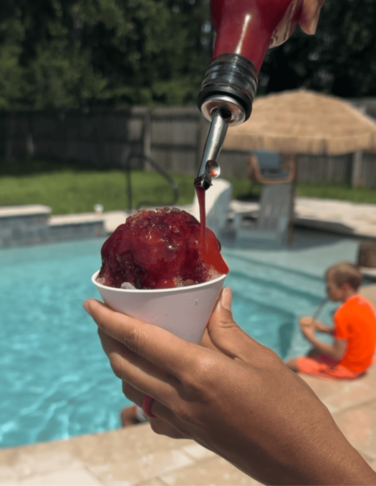 Pouring a mixed berry dye-free and refined sugar free snow cone syrup over shaved ice in a white snow cone cup. There is a little boy in an orange bathing suit and a pool in the background.