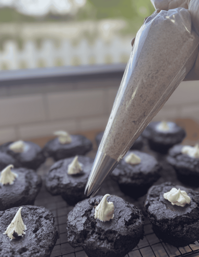 Hand piping cream onto freshly baked chocolate cupcakes with sprinkles.