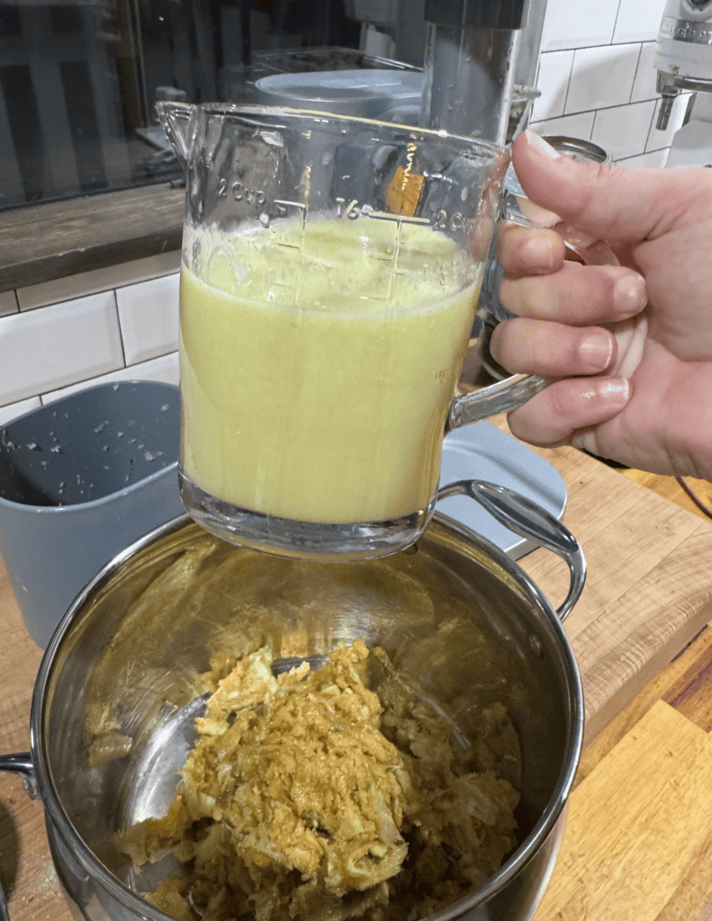 Fresh green juice being poured into a glass measuring cup, with a bowl of homemade dip on a wooden c.