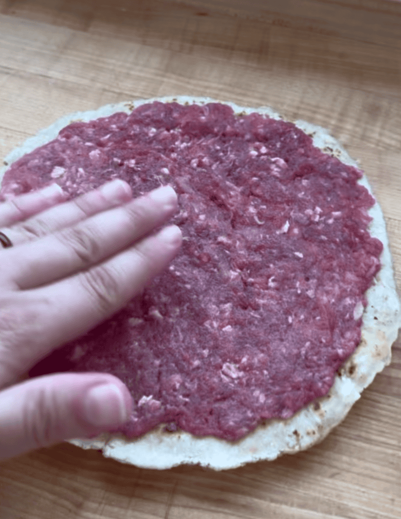 Hand spreading raw ground beef on pizza dough for homemade pizza.